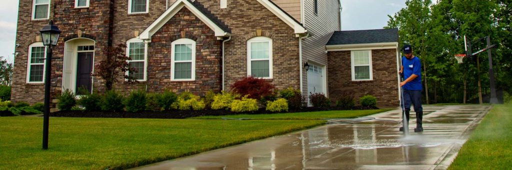 Power washing technician applying sealer to a concrete driveway in front of a brick home